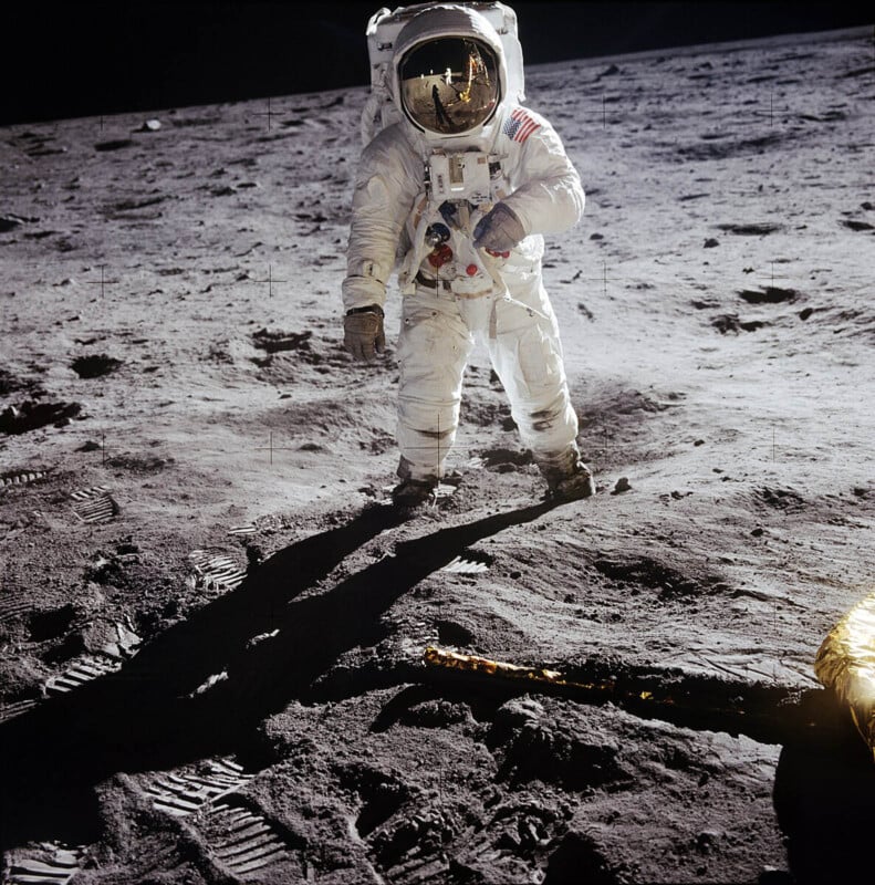Astronaut in a white spacesuit stands on the gray, dusty surface of the Moon, with boot prints visible. The astronaut’s visor reflects another person and the lunar lander. Shadows stretch across the ground.