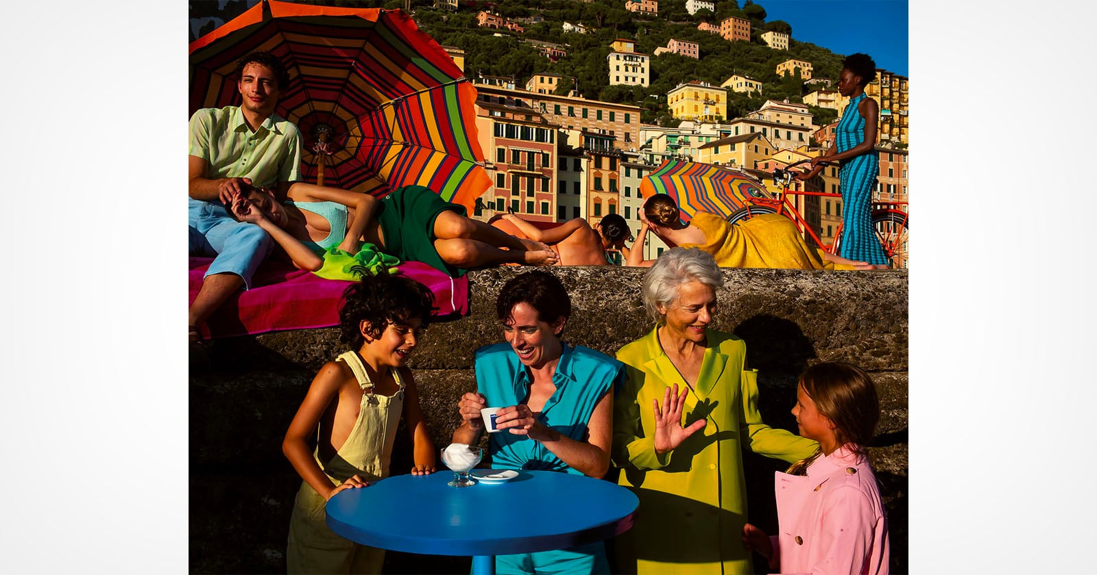 A group of people in colorful clothing socialize outdoors by a stone wall, with vibrant umbrellas and hillside houses in the background. Some sit at a blue table, while others relax or talk in the sunny, festive setting.