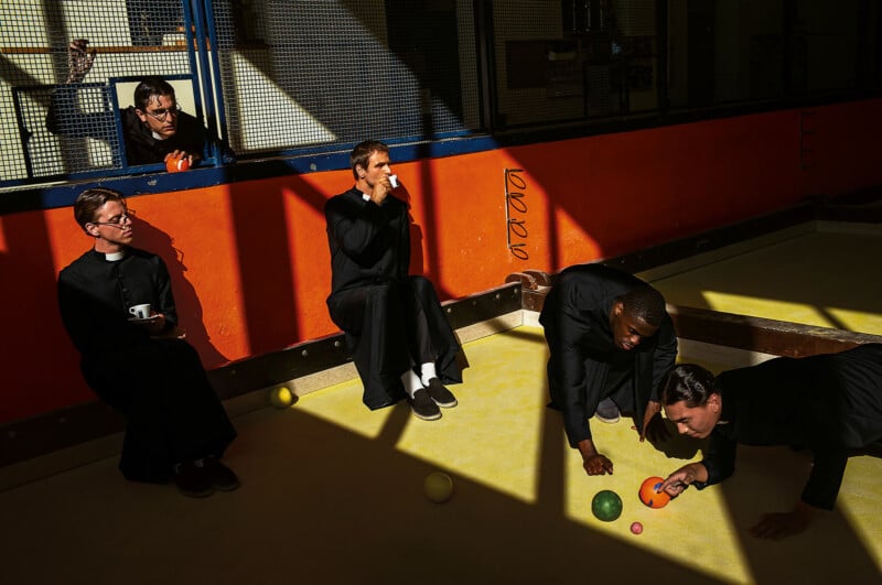 Five men in black robes, likely priests or seminarians, play bocce ball indoors on a yellow court. One sips from a cup, another watches from above behind a railing, while sunlight casts dramatic shadows across the scene.