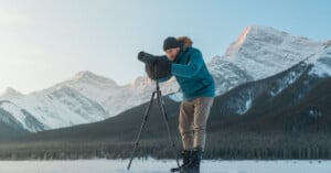 A person in winter clothing adjusts a camera on a tripod, standing on snow with snow-covered mountains and a clear sky in the background.
