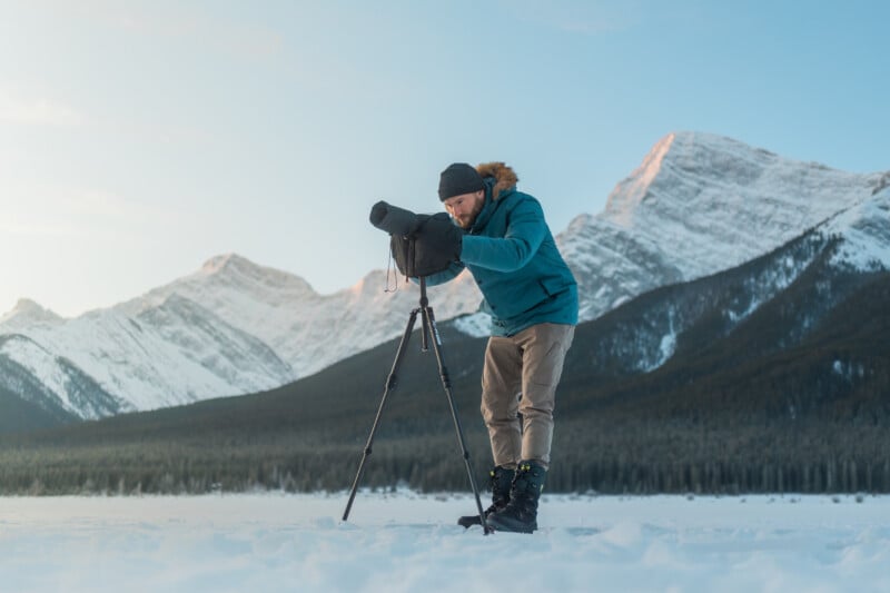 A person in winter clothing adjusts a camera on a tripod, standing on snow with snow-covered mountains and a clear sky in the background.