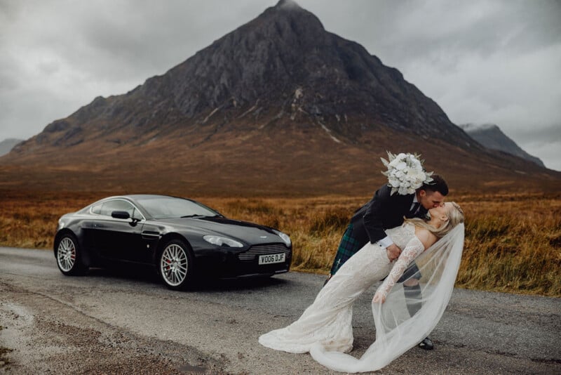 En una carretera rural, un novio con falda escocesa inclina la cabeza y besa a la novia delante de un coche deportivo negro. La novia sostiene un ramo y lleva un velo, con una majestuosa cadena montañosa al fondo.
