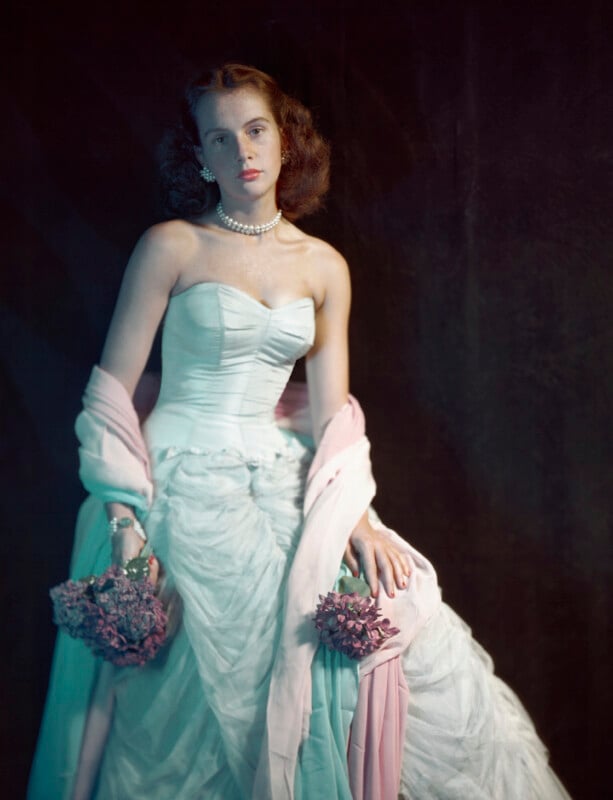 Una mujer joven viste un vestido blanco sin tirantes con un chal rosa sobre sus brazos y sostiene un ramo de flores rosas. Con un collar de perlas, mira suavemente hacia delante sobre un fondo oscuro.