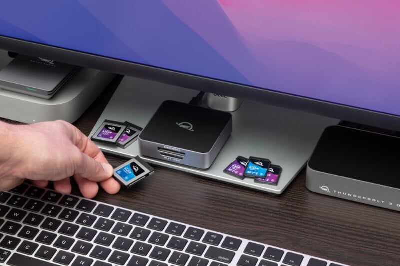 Una mano inserta una tarjeta de memoria en el lector de tarjetas debajo del monitor de la computadora, con varias otras tarjetas SD cerca y un teclado en primer plano.