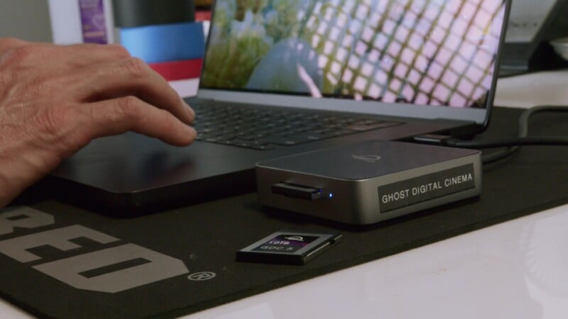 Una mano se acerca a una computadora portátil sobre una mesa junto a un cartel con la etiqueta "Cine digital fantasma" y una tarjeta SD colocada sobre una alfombrilla de ratón negra.