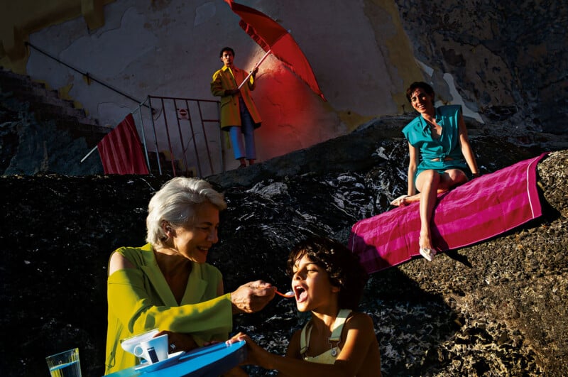 An older woman feeds a smiling child outdoors on rocky terrain, while a woman in teal sits on a pink towel above them. Another person stands in the background holding a red umbrella, with strong sunlight and shadows.