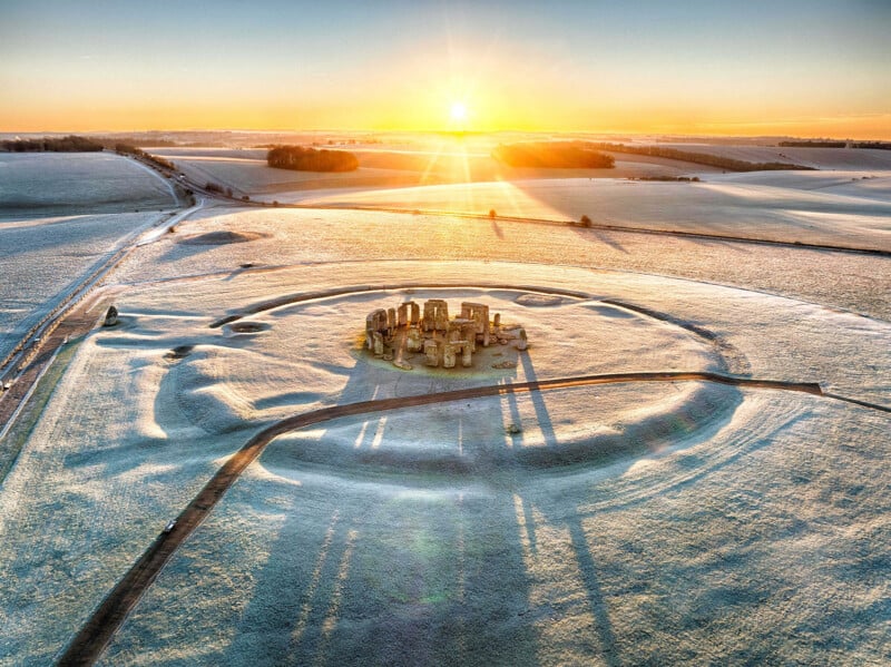 Aerial view of Stonehenge at sunrise, surrounded by frost-covered fields. The ancient stone circle casts long shadows, with the sun rising on the horizon and a clear sky overhead.