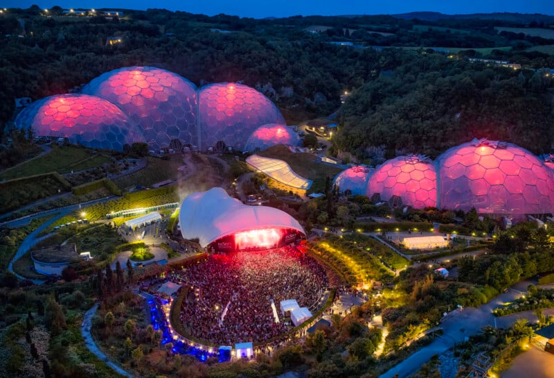 Aerial view of a large outdoor concert at night, with a crowd gathered in front of a stage, surrounded by illuminated geodesic domes glowing with pink and red lights, nestled in a lush, green landscape.