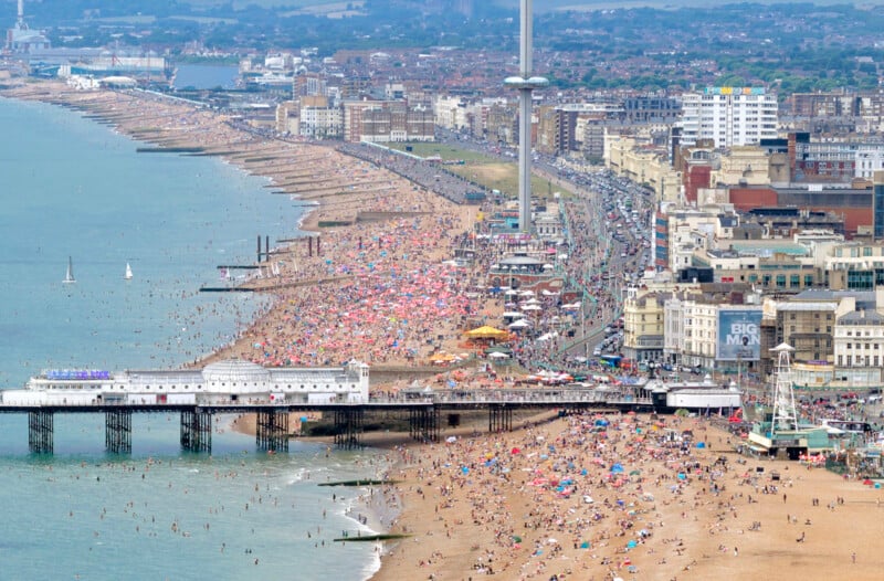 A crowded beach scene in Brighton, England, with people sunbathing and swimming. The pier extends into the sea, and the city’s buildings and the i360 observation tower are visible along the busy waterfront.
