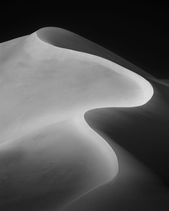 Fotografía en blanco y negro de suaves dunas de arena con curvas suaves y sombras dramáticas que crean un paisaje abstracto y minimalista contra un cielo oscuro.