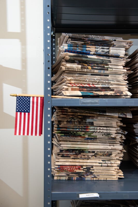 A small American flag is attached to a metal shelf stacked with several piles of folded newspapers. The newspapers are organized into two columns on the gray shelves.