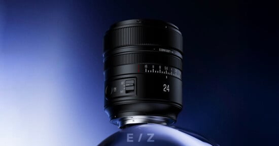 A black camera lens with "24" marked on it stands upright on a reflective, curved surface against a gradient blue and black background. The lens has focus and zoom rings, a switch, and engraved text.