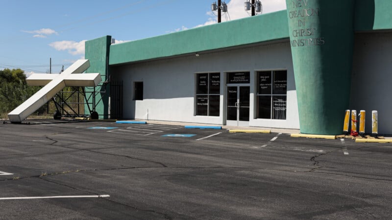 A large white cross has fallen and is leaning against a green and white church building. The parking lot is mostly empty, with a few handicapped spaces near the entrance. The sky is clear and sunny.
