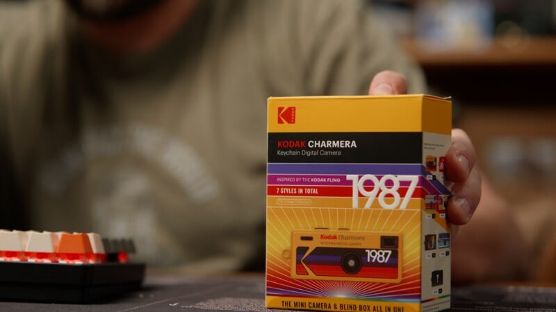 Una persona sostiene un colorido estuche para cámara digital con llavero Kodak Charmera, que está inspirado en la película Kodak de 1987 y presenta patrones retro y colores llamativos, con parte de un teclado visible en la mesa al lado.