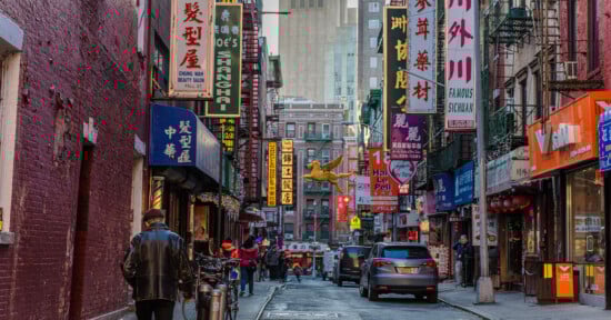 A busy street in Chinatown with colorful shop signs in Chinese, people walking, parked bikes, and cars. Tall buildings rise in the background, and a golden dragon decoration hangs above the street.