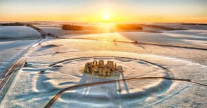 Aerial view of Stonehenge surrounded by frost-covered fields at sunrise, with long shadows from the stones and a golden sun on the horizon.