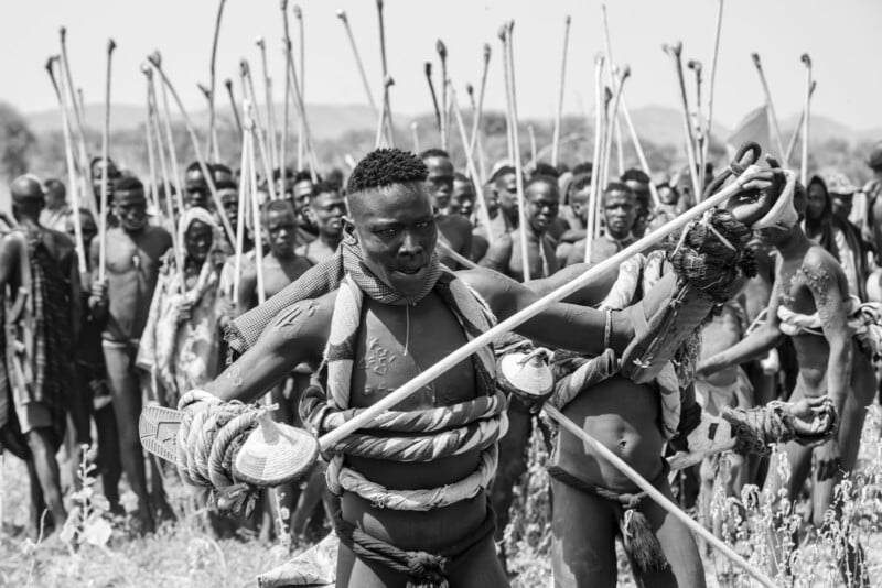 Un grupo de hombres, algunos con pintura corporal y cordones decorativos, se encuentran al aire libre sosteniendo palos largos o bastones, con un hombre al frente haciendo gestos dinámicos. La escena parece ser ritualista o tradicional.
