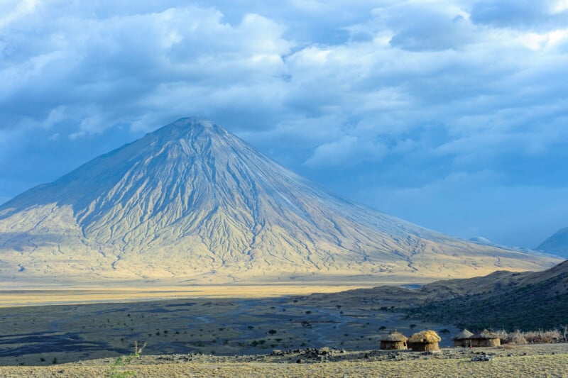Bajo un cielo nublado, un imponente pico cónico se alza sobre una vasta llanura seca, con varias pequeñas cabañas con techo de paja agrupadas en primer plano.