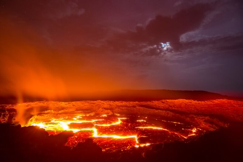 Por la noche, la lava dentro del cráter activo brilla de color naranja y rojo brillante, el vapor se eleva y las nubes oscurecen parte de la luna en un cielo oscuro y dramático.