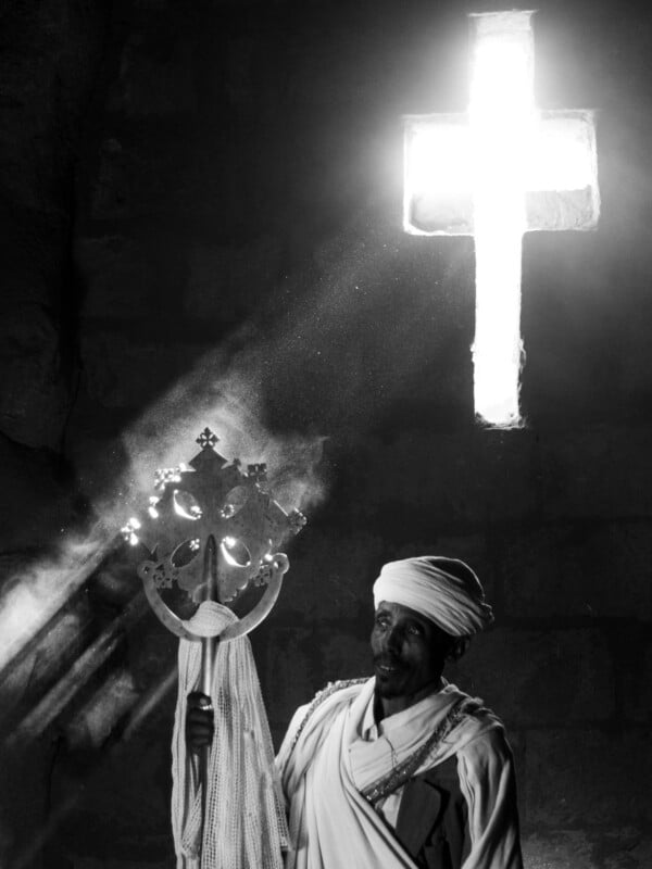 Un hombre vestido con ropa tradicional etíope sostiene una cruz ornamentada y se para frente a un muro de piedra mientras la luz del sol brilla a través de una ventana brillante en forma de cruz arriba. La imagen está en blanco y negro.