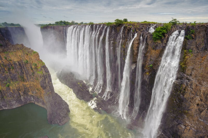 Una amplia y espectacular cascada cae desde un acantilado rocoso, rodeada de exuberante vegetación verde, bajo un cielo nublado, creando niebla en la base donde el agua desemboca en el río.