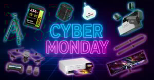 A neon "Cyber Monday" sign glows at the center, surrounded by electronics like a camera tripod, SD card, printer, gaming monitor, camera bag, drone, batteries, chargers, and tech accessories on a dark grid background.