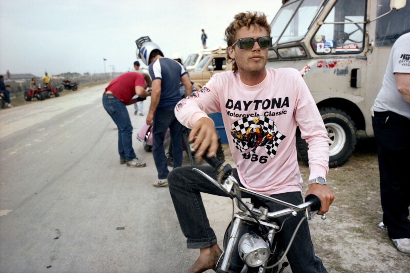 A man wearing sunglasses and a pink "Daytona Motorcycle Classic 1986" shirt sits on a small motorcycle. Behind him, several people stand near vans and motorbikes in an outdoor setting.