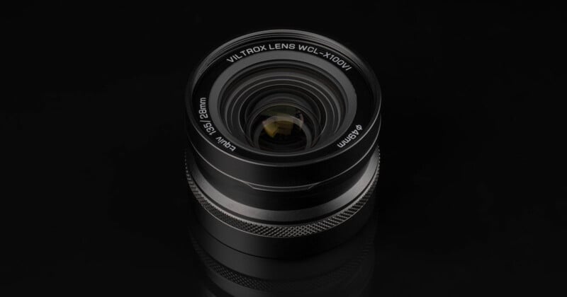 Vista cercana de una lente de cámara Viltrox de 28 mm con rosca de filtro de 49 mm, colocada sobre una superficie reflectante negra. La toma y sus detalles están nítidamente enfocados.