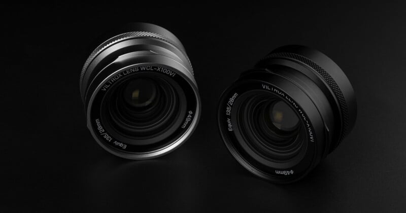 Dos lentes de cámara con acabados metálicos (uno plateado y otro negro) se colocan uno al lado del otro sobre una superficie oscura. Las marcas y reflejos de la lente son claramente visibles, resaltando los detalles de su diseño.