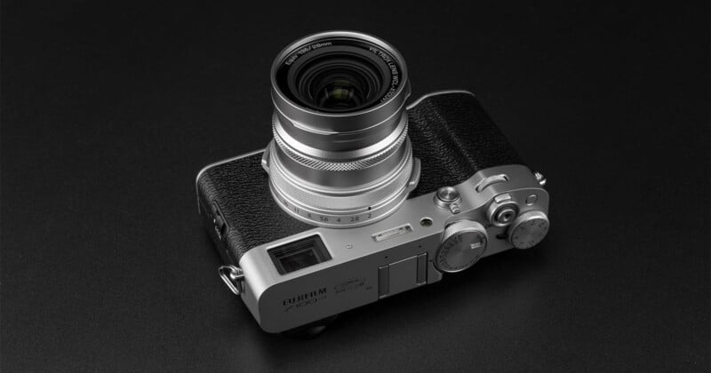 Una cámara Fujifilm X100VI plateada y negra con una lente grande descansa sobre una superficie oscura y brillante. La cámara presenta diales y botones en la parte superior y un agarre texturizado en el lado derecho.