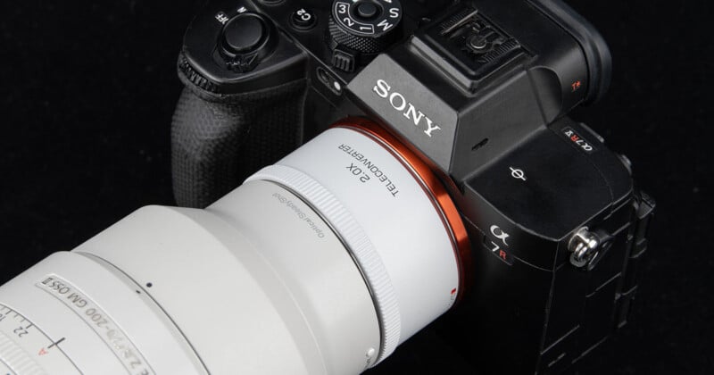 Primer plano de una cámara Sony α7R negra con un gran teleobjetivo blanco y un teleconvertidor de 2,0 aumentos sobre un fondo oscuro.