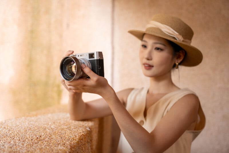 Una mujer con un vestido beige y un sombrero de paja sostiene una cámara antigua y se prepara para tomar fotografías. Está parada junto a una pared texturizada bajo una suave luz natural, mirando fijamente a la cámara.