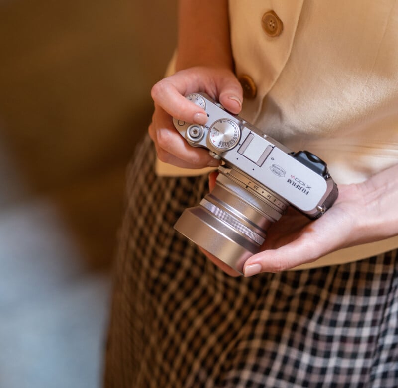 Un hombre con una camisa color crema y una falda a cuadros sostiene una cámara Fuji plateada y ajusta el dial con una mano. La escena está suavemente iluminada y la atención se centra en la cámara y las manos.