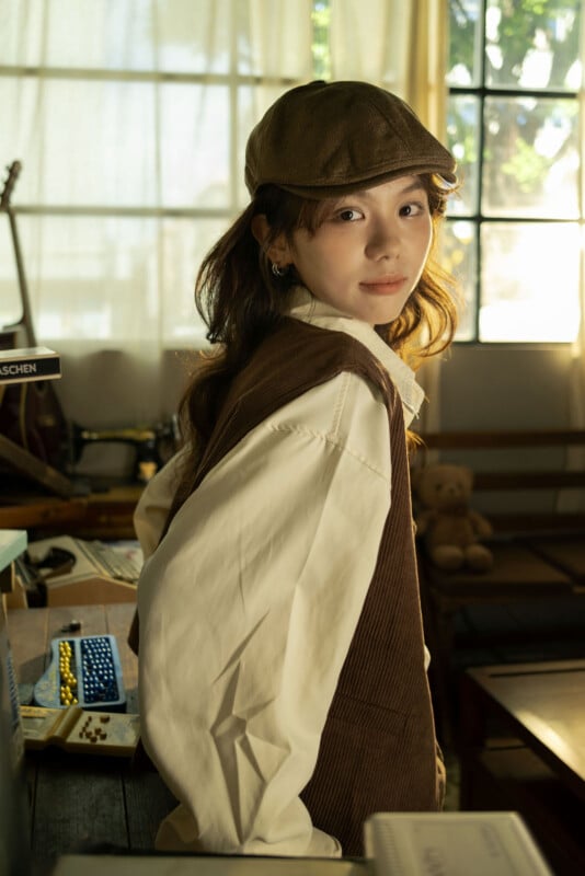 Un joven con camisa blanca, sombrero marrón y chaleco está apoyado en una mesa en una habitación soleada con grandes ventanales; al fondo se ve un osito de peluche, una guitarra y libros.