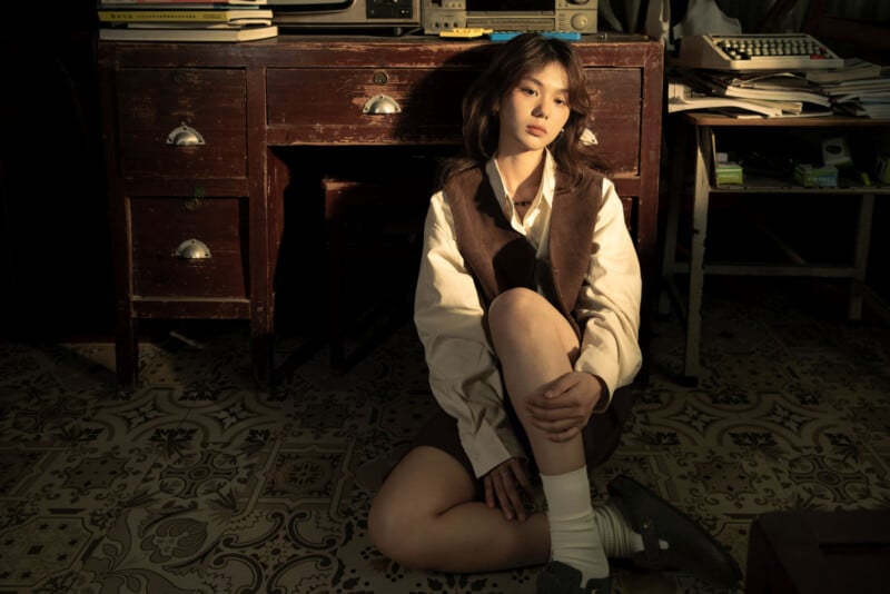 Una mujer joven se sienta en un suelo estampado frente a una mesa de madera con un brazo sobre las rodillas. Llevaba una blusa blanca, un chaleco y una falda marrones, y el sol proyectaba sombras sobre y alrededor de su rostro.