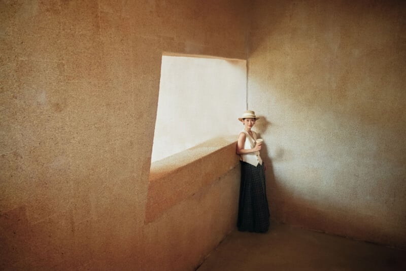 En una habitación sencilla y de textura beige, un hombre con sombrero, chaleco blanco y una falda larga y oscura se encuentra junto a una ventana grande y angular, dejando entrar una luz suave.