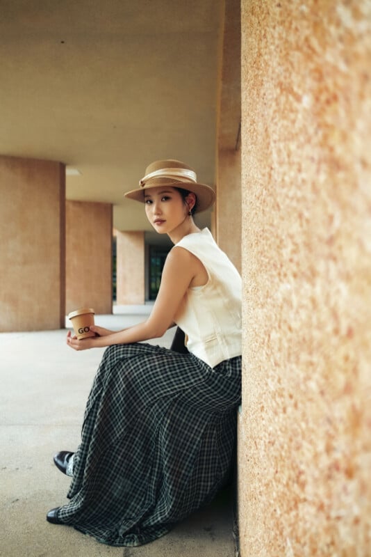 Una mujer con sombrero y camiseta sin mangas está sentada contra una pared texturizada, sosteniendo una taza de café. Con una falda larga a cuadros, miró a la cámara en un escenario al aire libre con columnas altas.
