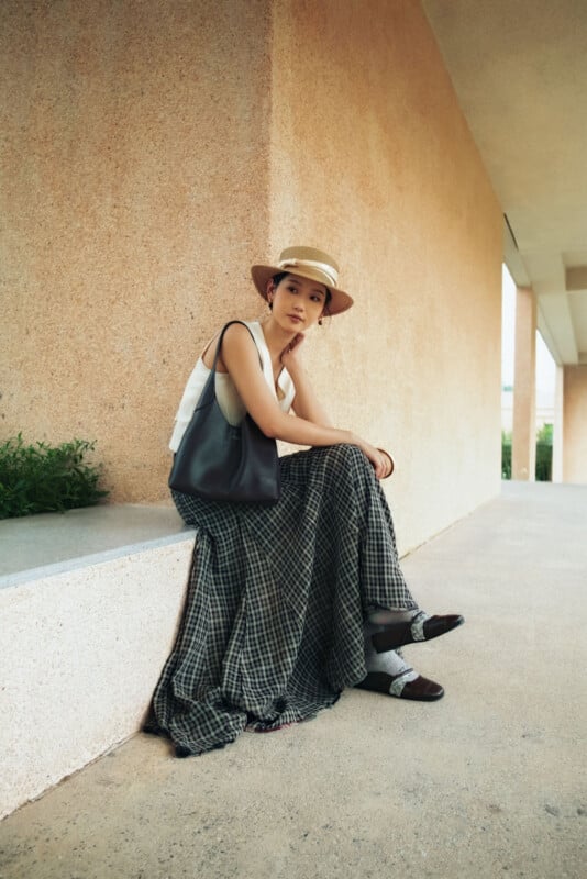 Una mujer que lleva un sombrero de paja, una blusa sin mangas y una falda a cuadros está sentada en una repisa baja de concreto afuera del edificio. Llevaba un bolso negro y miraba pensativamente a la cámara.