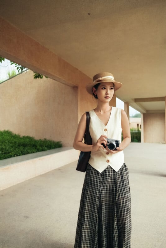 Una mujer con sombrero de paja, camiseta color crema y falda a cuadros está parada al aire libre con una cámara en la mano, sobre un fondo de plantas verdes y un muro de hormigón beige.