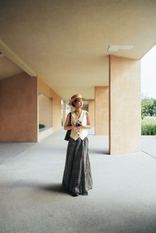 Un hombre con sombrero, chaleco color crema y pantalones anchos a cuadros estaba parado debajo de la pasarela cubierta, mirando hacia otro lado con una cámara. El fondo incluye paredes de color melocotón y plantas verdes.