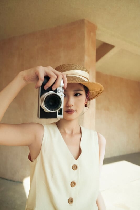 Una mujer que lleva una blusa color crema sin mangas y un sombrero de paja sostiene una cámara frente a su cara como si estuviera tomando una fotografía, sobre un fondo de paredes beige y luz solar.