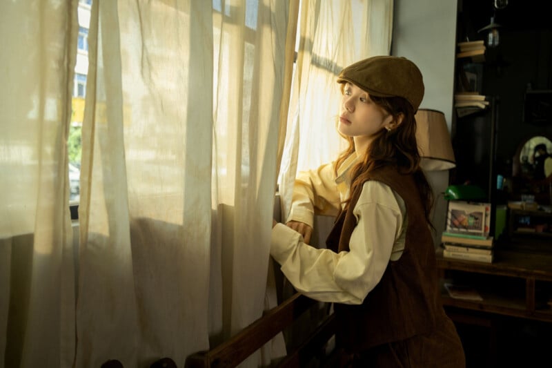 Una mujer joven con ropa de estilo retro y un sombrero marrón se encuentra junto a la ventana y mira a través de las cortinas transparentes. La cálida luz del sol entraba iluminando su expresión pensativa.