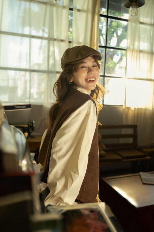 En una habitación soleada con grandes ventanales y decoración vintage, una mujer vestida con camisa blanca, sombrero marrón y chaleco se gira hacia la cámara con una sonrisa.