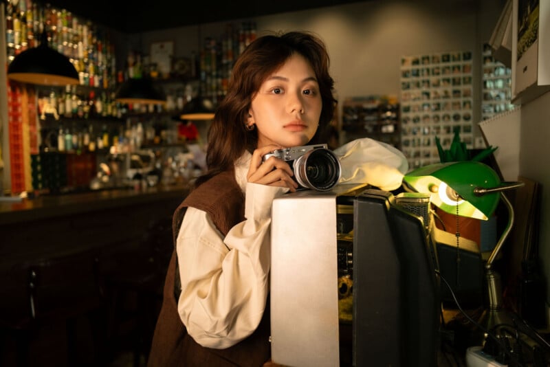 Un hombre con el pelo hasta los hombros sostiene una cámara y se apoya en un viejo televisor en una acogedora habitación con poca luz, llena de botellas y fotografías, iluminada por una lámpara de escritorio verde.