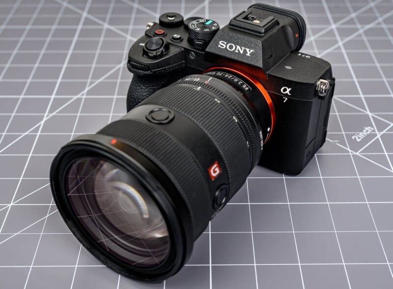 A Sony Alpha mirrorless camera with a large zoom lens is placed on a gray, grid-patterned surface. The camera body is black with visible buttons and dials.