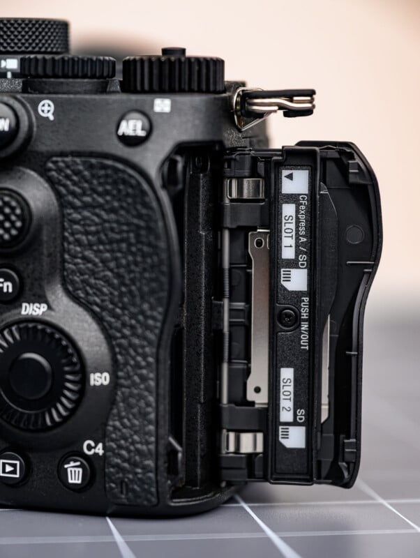 Close-up of a digital camera with its side compartment open, revealing two SD card slots labeled “SLOT 1” and “SLOT 2.” The camera body shows various buttons and textured grip details.