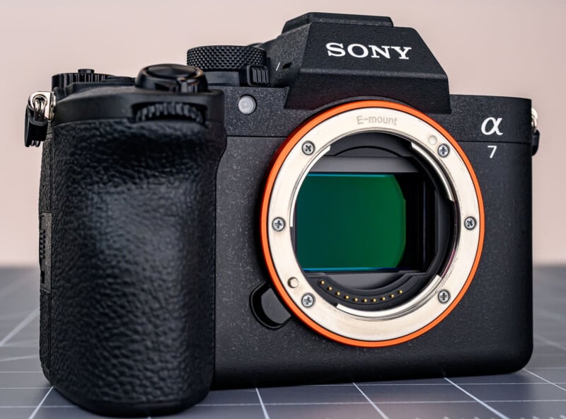 A close-up of a Sony Alpha 7 mirrorless camera body without a lens, showing the E-mount and image sensor. The camera is black with a textured grip and is placed on a gray, grid-patterned surface.