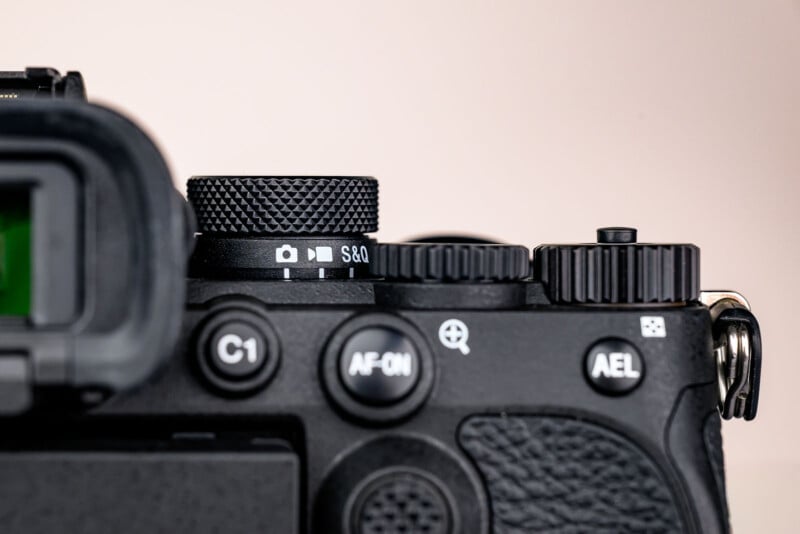 Close-up of a digital camera's control dials and buttons, including the mode dial with icons, an AF-ON button, C1 button, and AEL button, with a textured black surface and part of the viewfinder visible.