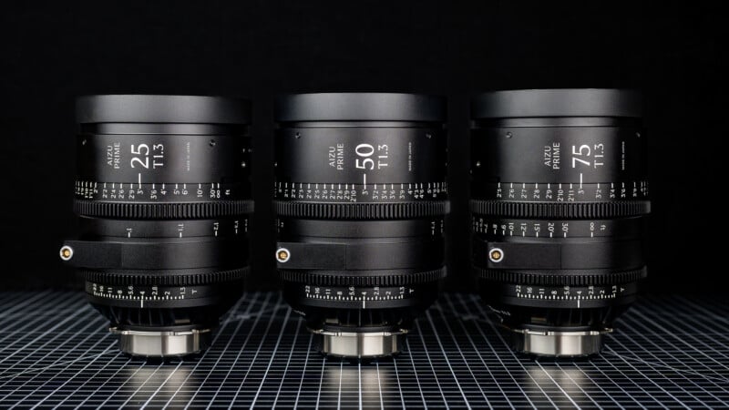 Tres lentes de cámara profesional con etiquetas de 25 mm, 50 mm y 75 mm están dispuestas una al lado de la otra sobre una superficie a cuadros sobre un fondo negro, mostrando sus marcas detalladas de escala de apertura y enfoque.