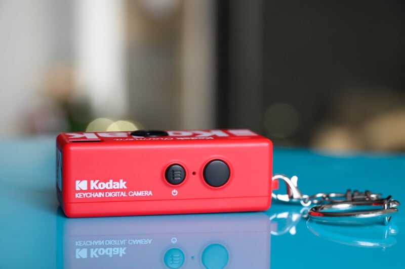 Una pequeña cámara digital con llavero Kodak roja se encuentra sobre una superficie azul brillante con un llavero de metal adjunto al costado. La cámara tiene botones y marca en el lado visible.